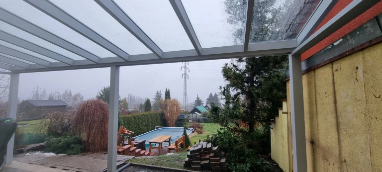 Widok z aluminiowej pergoli na ogród z basenem w deszczowy dzień