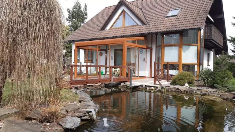Zaszklona pergola z aluminium przy domu jednorodzinnym z oczkiem wodnym i wierzbą.