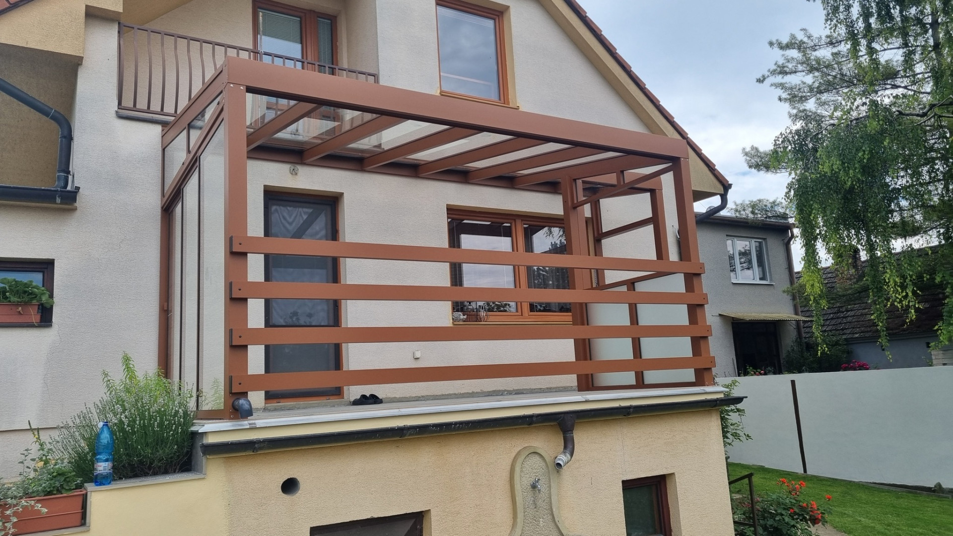 Brązowa, nietypowa pergola aluminiowa ze szklanym dachem i bocznymi osłonami przeciwsłonecznymi na balkonie domu jednorodzinnego
