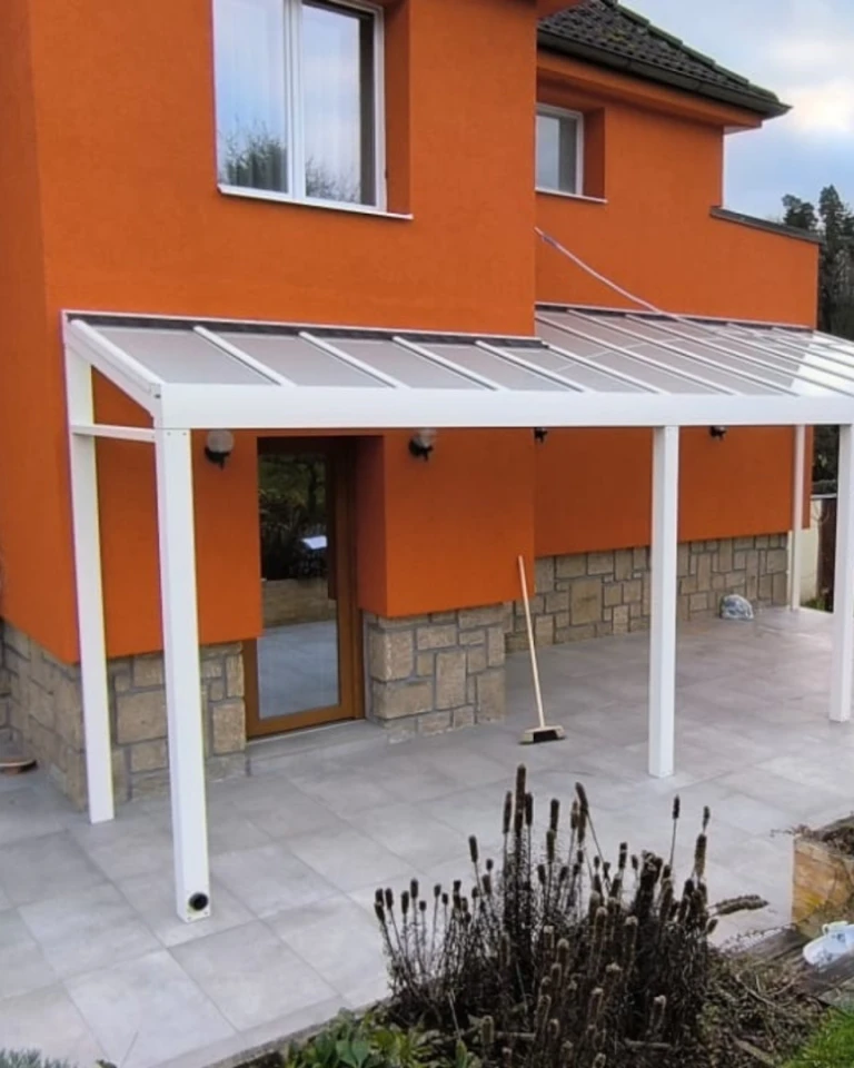 Nowoczesna biała pergola z aluminium przymocowana do pomarańczowego domu ze szklanym dachem i kostką brukową
