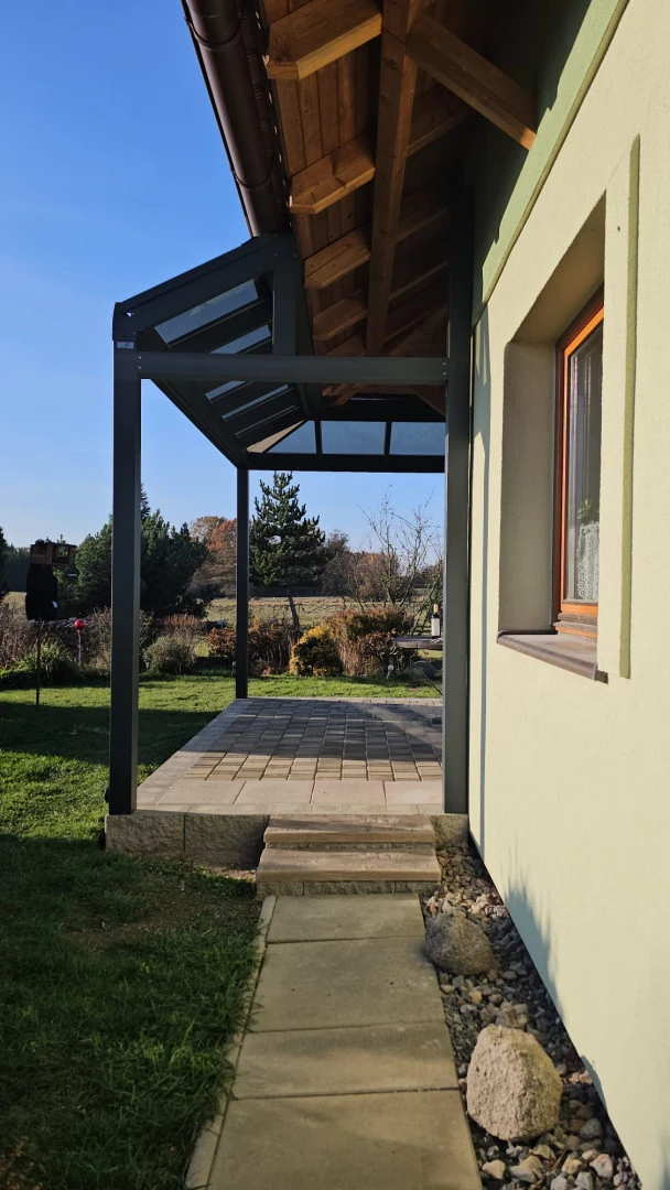 Widok z boku na nowoczesną aluminiową pergolę z przeszklonym dachem, przylegającą do domu jednorodzinnego z widokiem na ogród.