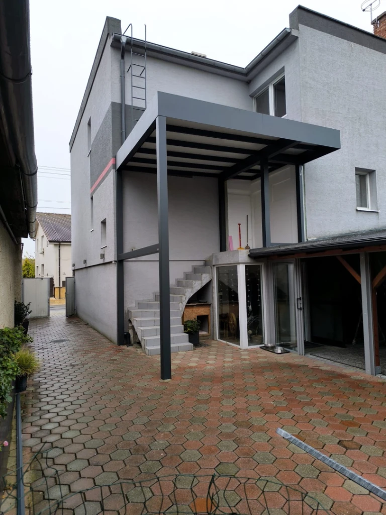 Nowoczesna, ciemnoszara pergola aluminiowa ze stałym zadaszeniem, przymocowana do elewacji domu jednorodzinnego nad zewnętrznymi schodami.