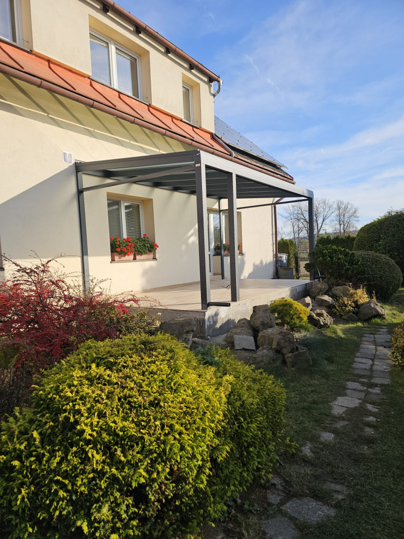 Nowoczesna pergola aluminiowa z przeszklonym dachem, dobudowana do domu jednorodzinnego z ogrodem.