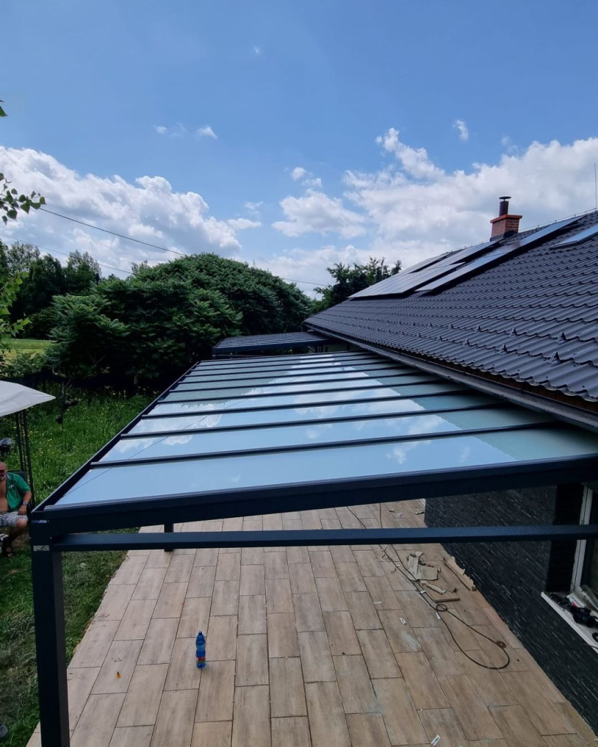 Nowoczesna pergola aluminiowa ze szklanym dachem, dobudowana do domu jednorodzinnego z widokiem na ogród.