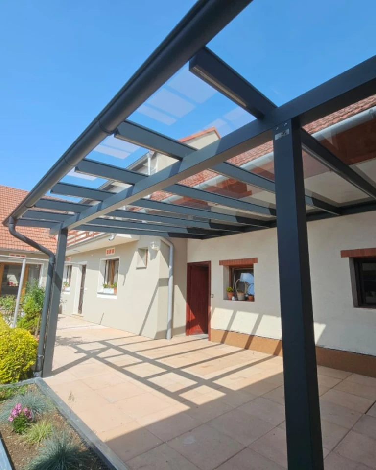 Nowoczesna, antracytowa pergola z aluminium ze szklanym dachem, dobudowana do domu jednorodzinnego z wybrukowanym tarasem.