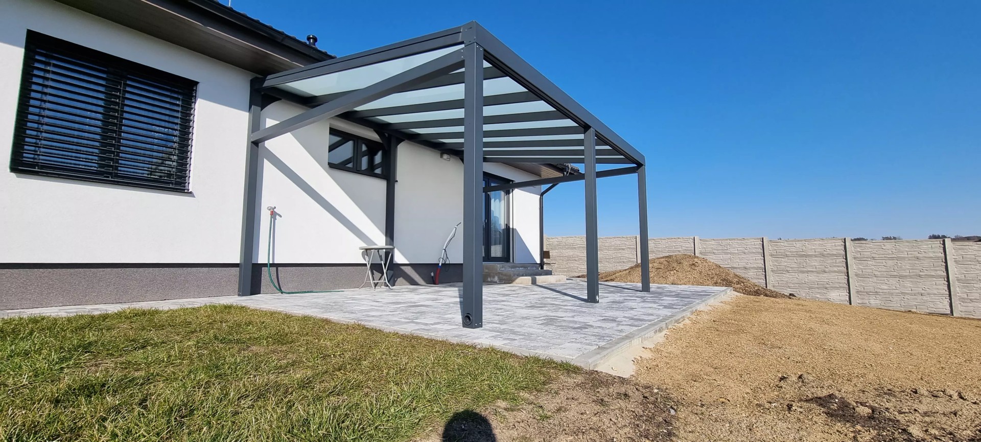 Nowoczesna antracytowa pergola z aluminium ze szklanym dachem, dobudowana do domu jednorodzinnego na wyłożonym płytkami tarasie.