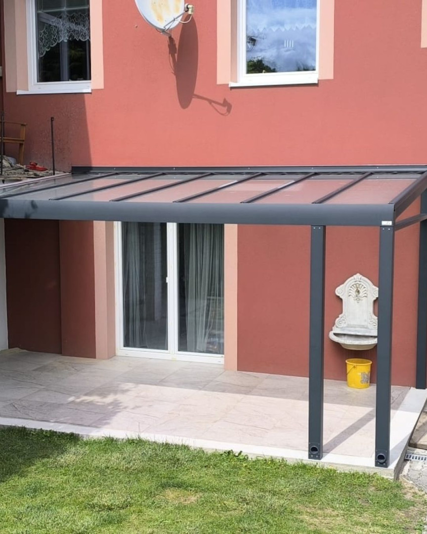 Nowoczesna pergola aluminiowa z przeszklonym dachem, zamontowana przy ścianie czerwonego domu nad tarasem.
