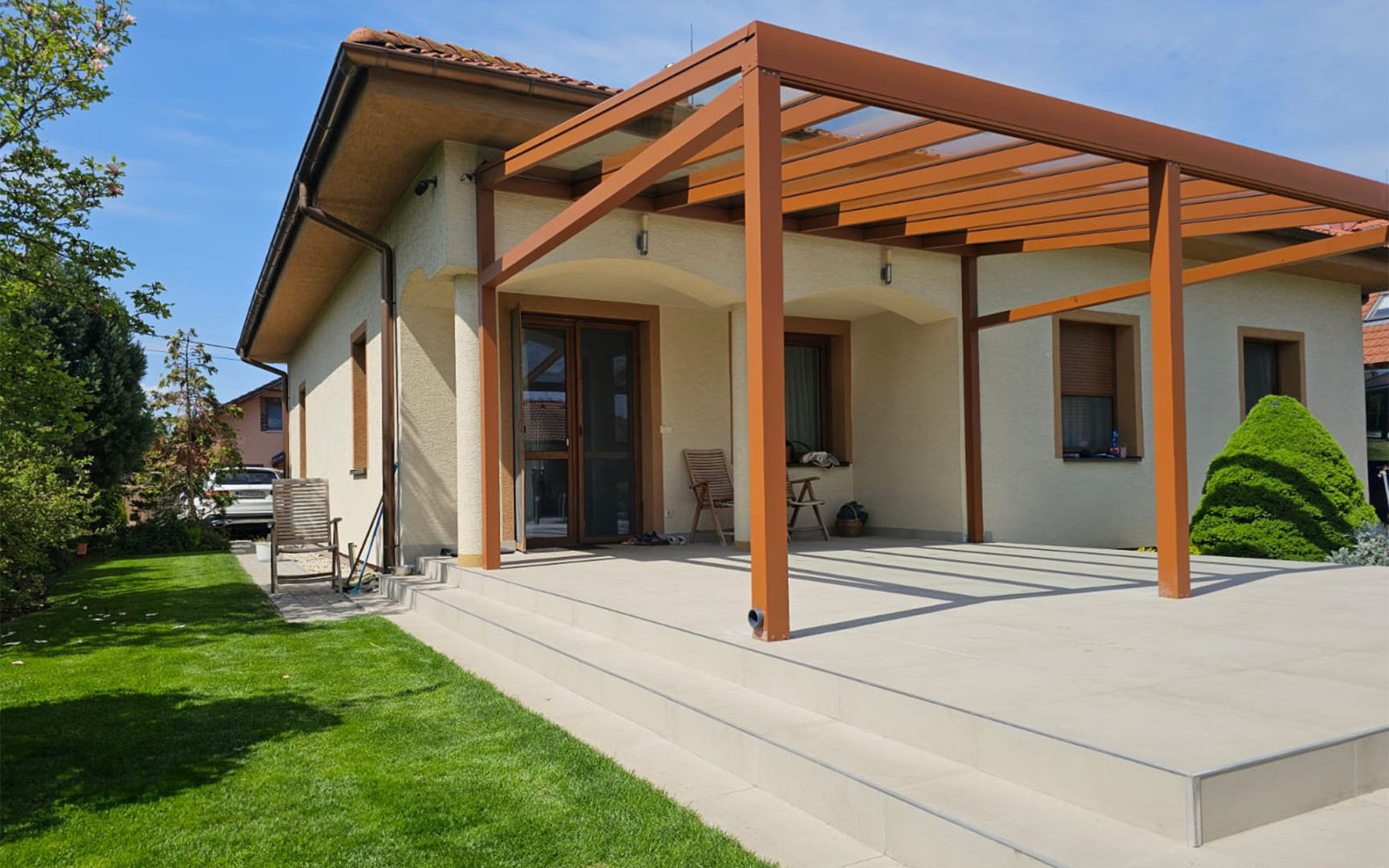 Nowoczesna pergola z aluminium w kolorze brązowym, z przeszklonym dachem, dobudowana do domu jednorodzinnego z przestronnym tarasem.