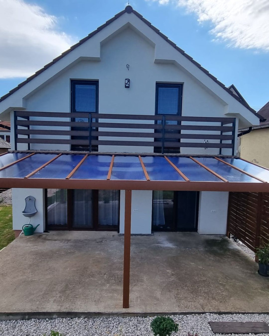 Brązowa pergola z aluminium ze szklanym dachem dobudowana do domu jednorodzinnego z balkonem.