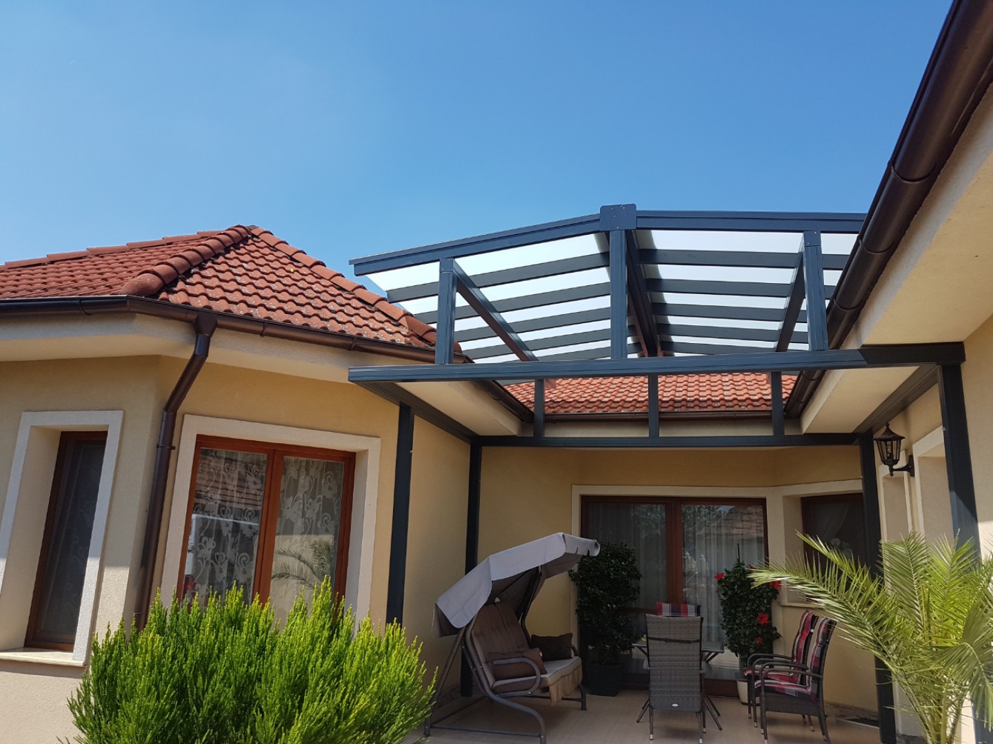 Nowoczesna aluminiowa pergola ROBUST z przeszklonym dachem, dobudowana do domu jednorodzinnego, z meblami tarasowymi i huśtawką.