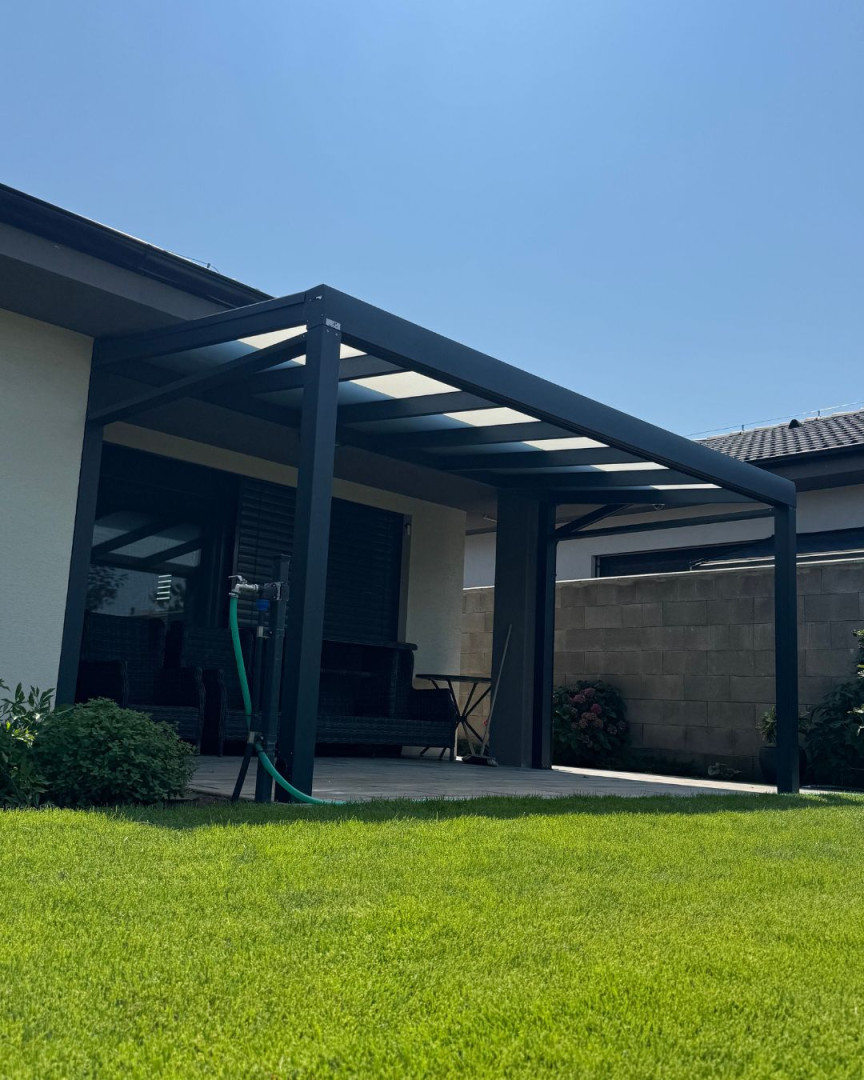 Nowoczesna, ciemnoszara pergola aluminiowa przylegająca do domu, ze szklanym dachem i wypielęgnowanym trawnikiem