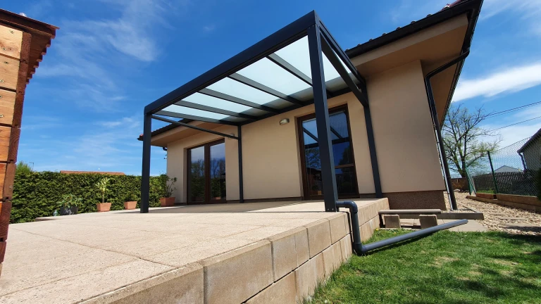 Nowoczesna, ciemnoszara pergola z aluminium ze szklanym dachem, zamontowana przy domu jednorodzinnym na wybrukowanym tarasie.