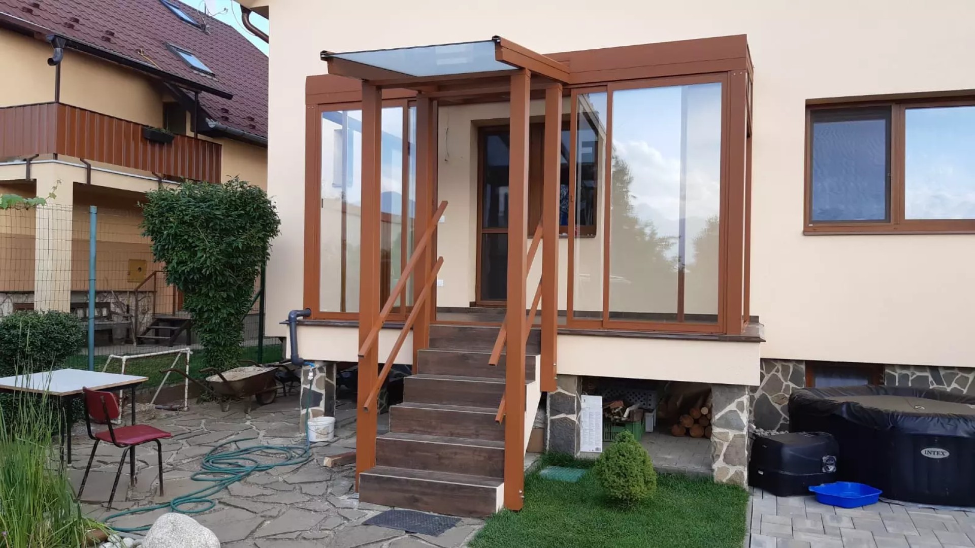 Brązowa, przeszklona pergola z aluminium z zadaszeniem nad schodami wejściowymi przy domu jednorodzinnym.