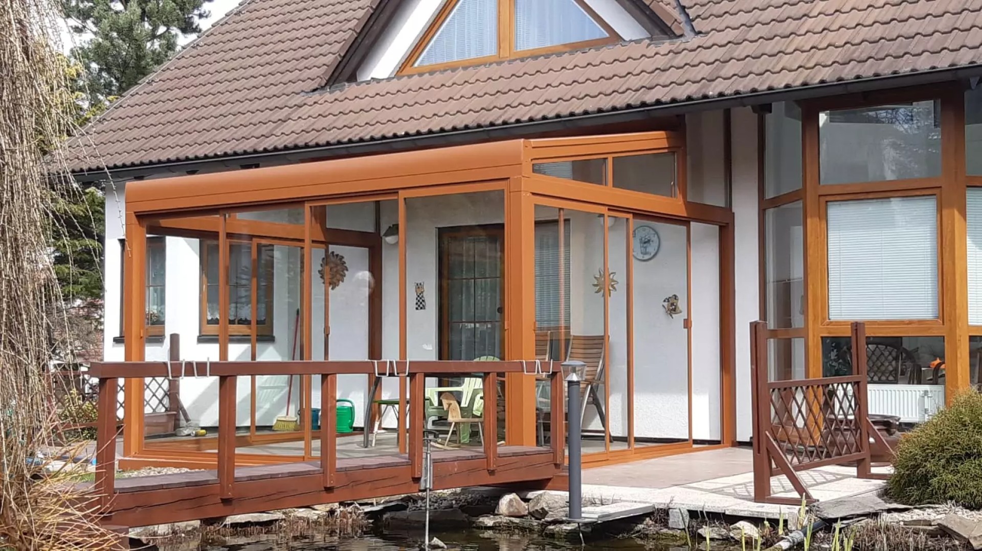 Zaszklona pergola aluminiowa ROBUST w kolorze brązowym przy domu jednorodzinnym z drewnianym mostkiem
