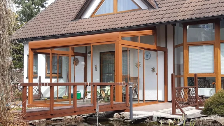 Zaszklona pergola aluminiowa ROBUST w kolorze brązowym przy domu jednorodzinnym z drewnianym mostkiem