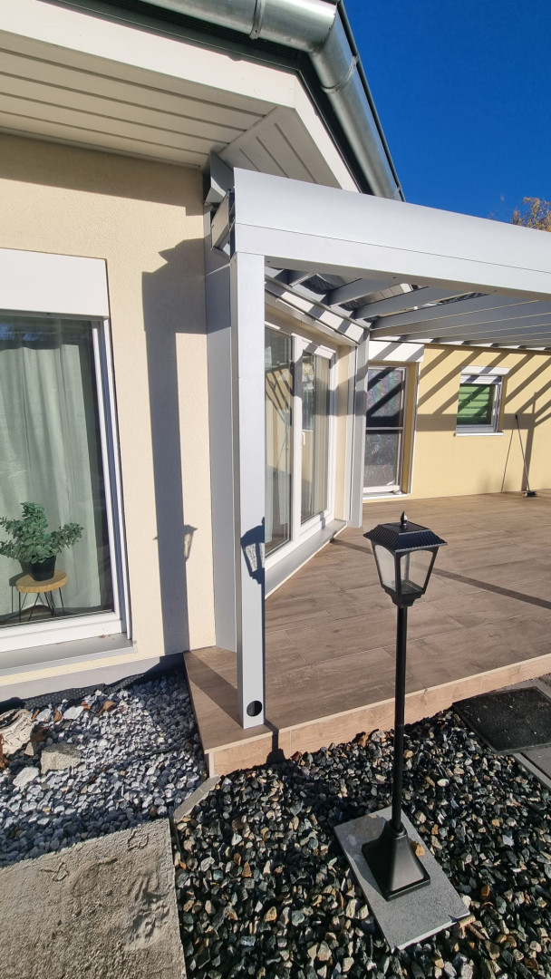 Szczegół mocowania białej aluminiowej pergoli do elewacji domu na tarasie z drewnianymi elementami dekoracyjnymi.