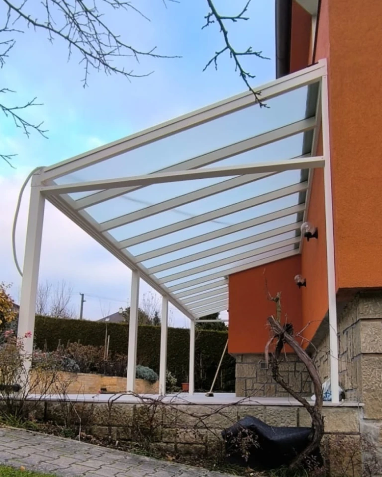 Nowoczesna biała pergola z aluminium ze szklanym dachem, przymocowana do pomarańczowego domu jednorodzinnego