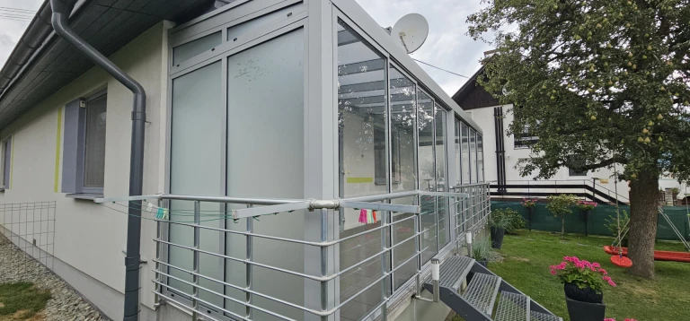 Nowoczesny, przeszklony ogród zimowy przylegający do domu jednorodzinnego, z klatką schodową i balustradą ze stali nierdzewnej.