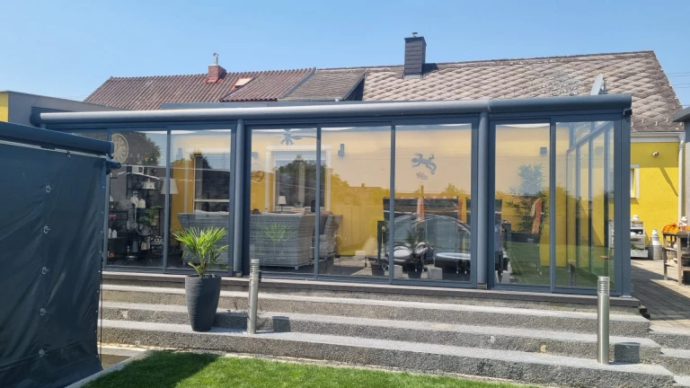 Nowoczesna, przeszklona, aluminiowa oranżeria w kolorze antracytowym, dobudowana do żółtego domu jednorodzinnego z klatką schodową.