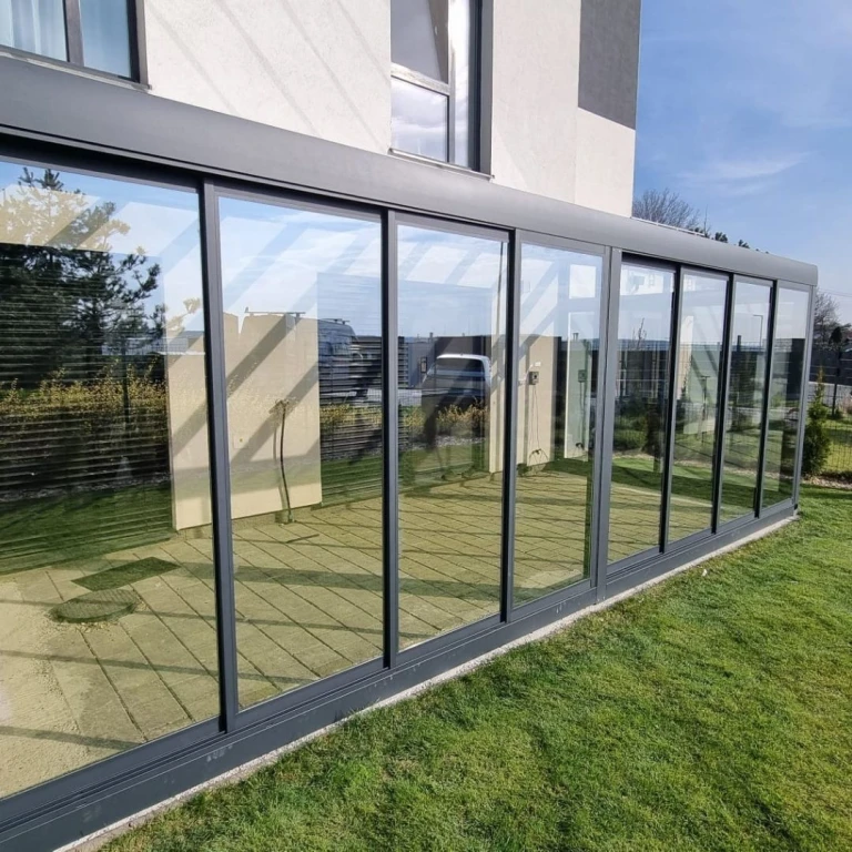 Nowoczesna, przeszklona konstrukcja ogrodu zimowego z aluminium, dobudowana do domu jednorodzinnego z zielonym trawnikiem.