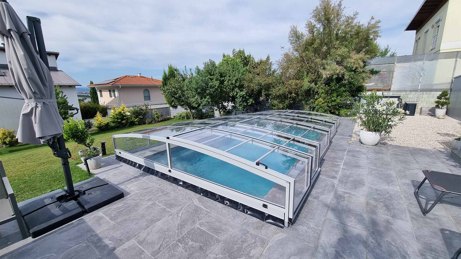 Nowoczesna, niska aluminiowa konstrukcja zadaszenia basenu PREMIER na wyłożonym płytkami tarasie w ogrodzie domu jednorodzinnego