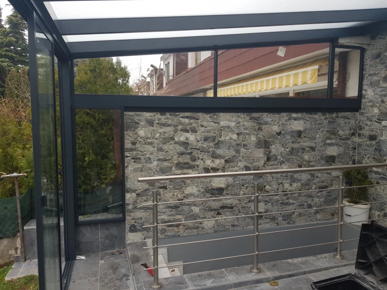 Nowoczesny ogród zimowy z antracytową konstrukcją aluminiową i kamienną okładziną na ścianie.