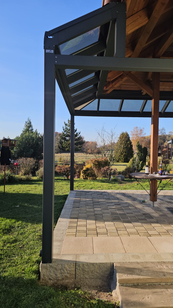 Szczegółowe ujęcie aluminiowej konstrukcji i przeszklonego dachu pergoli z widokiem na ogród.