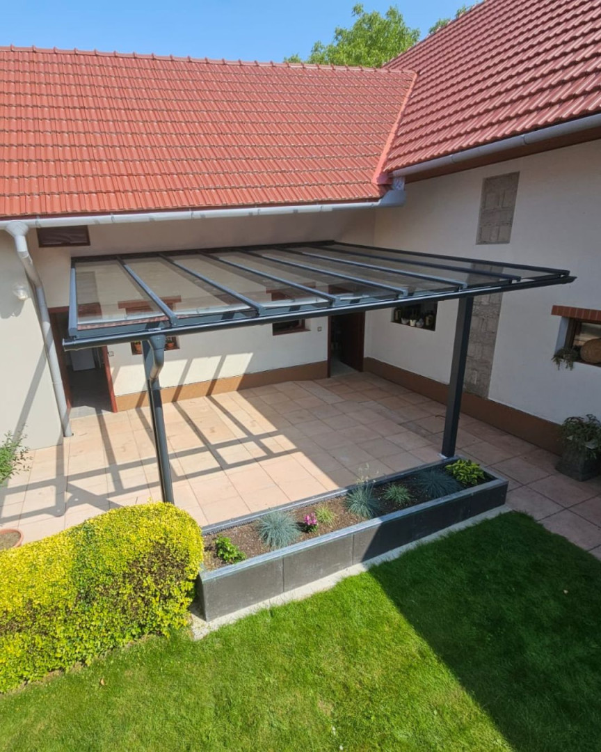 Nowoczesna pergola aluminiowa ze szklanym dachem, przymocowana do ściany domu z wyłożonym płytkami tarasem.