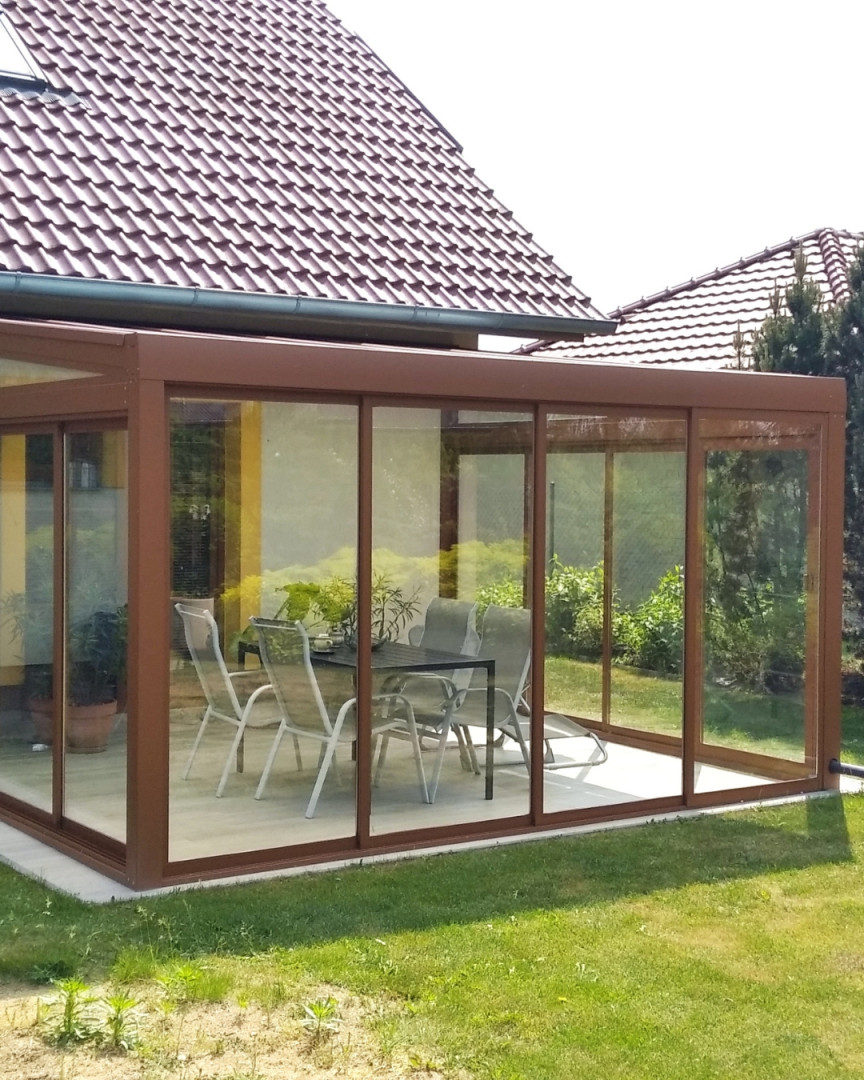 Zaszklona aluminiowa oranżeria z brązową ramą i meblami ogrodowymi na tarasie przy domu