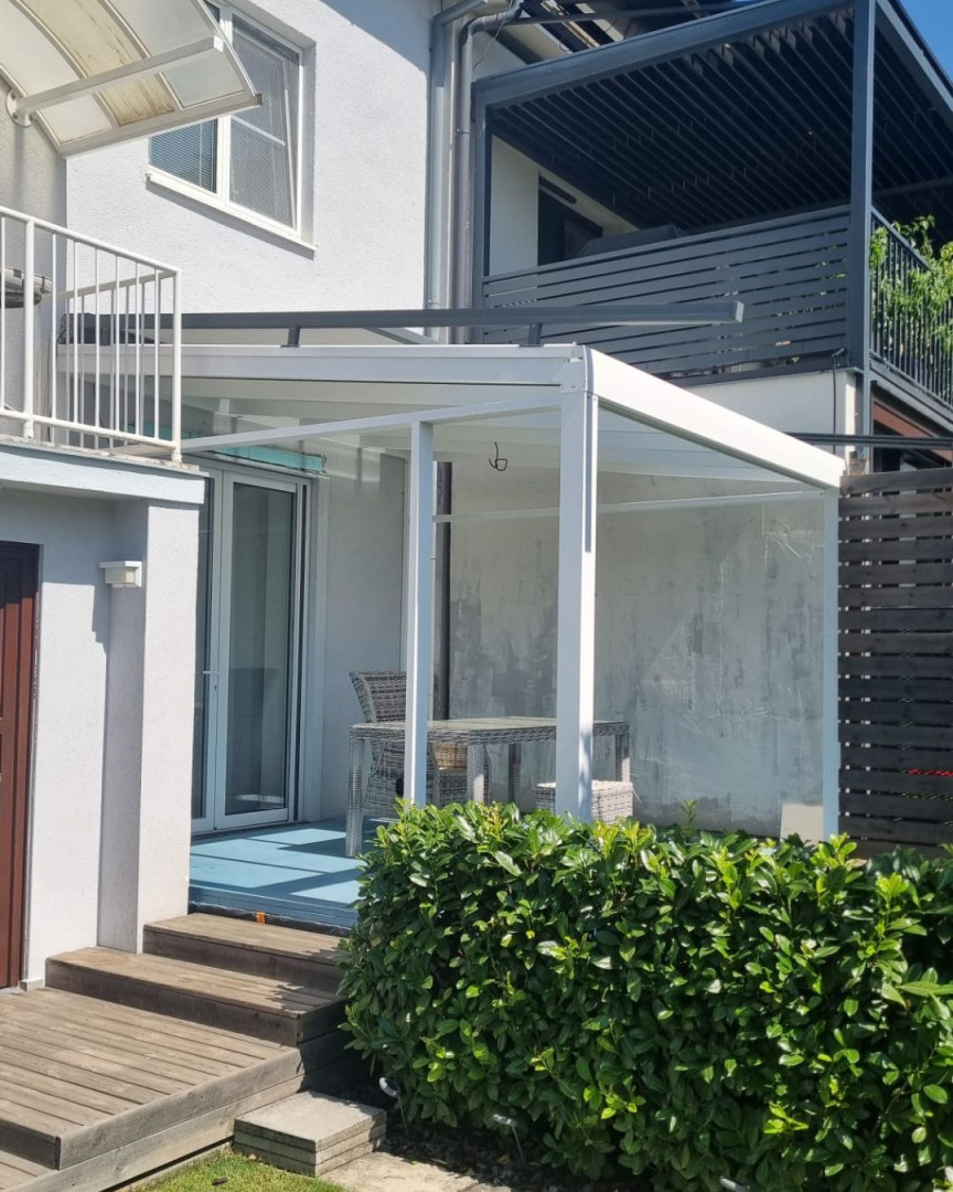 Biała, nowoczesna pergola z aluminium ze szklanym dachem przy domu jednorodzinnym z meblami tarasowymi.