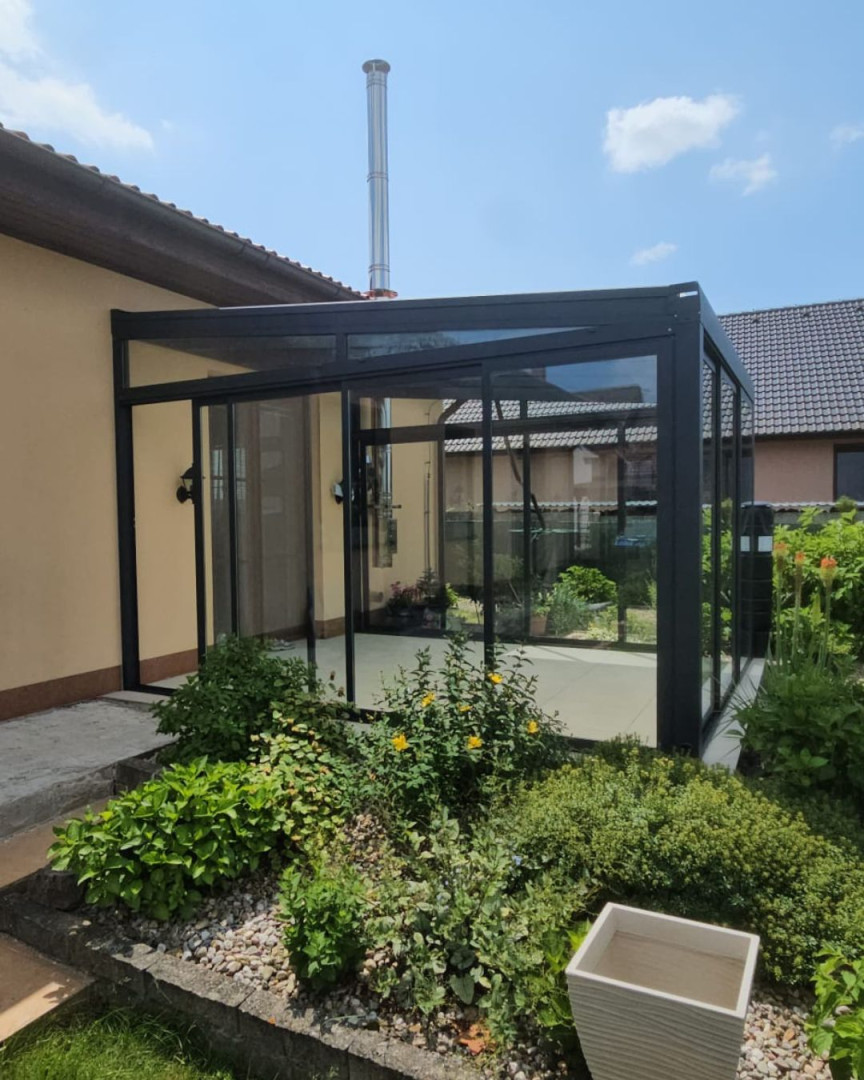 Nowoczesna, przeszklona dobudówka do domu jednorodzinnego z czarną ramą i drzwiami przesuwnymi, otoczona ogrodem.