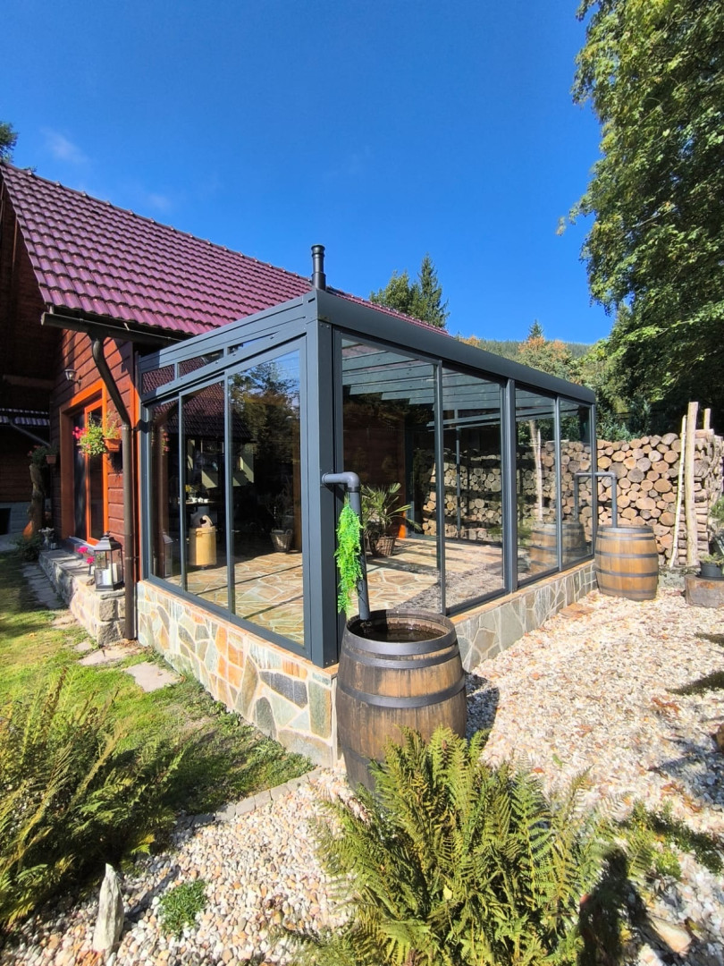 Nowoczesna, przeszklona oranżeria z ciemną aluminiową ramą, dobudowana do drewnianego domu z kamiennym cokołem.