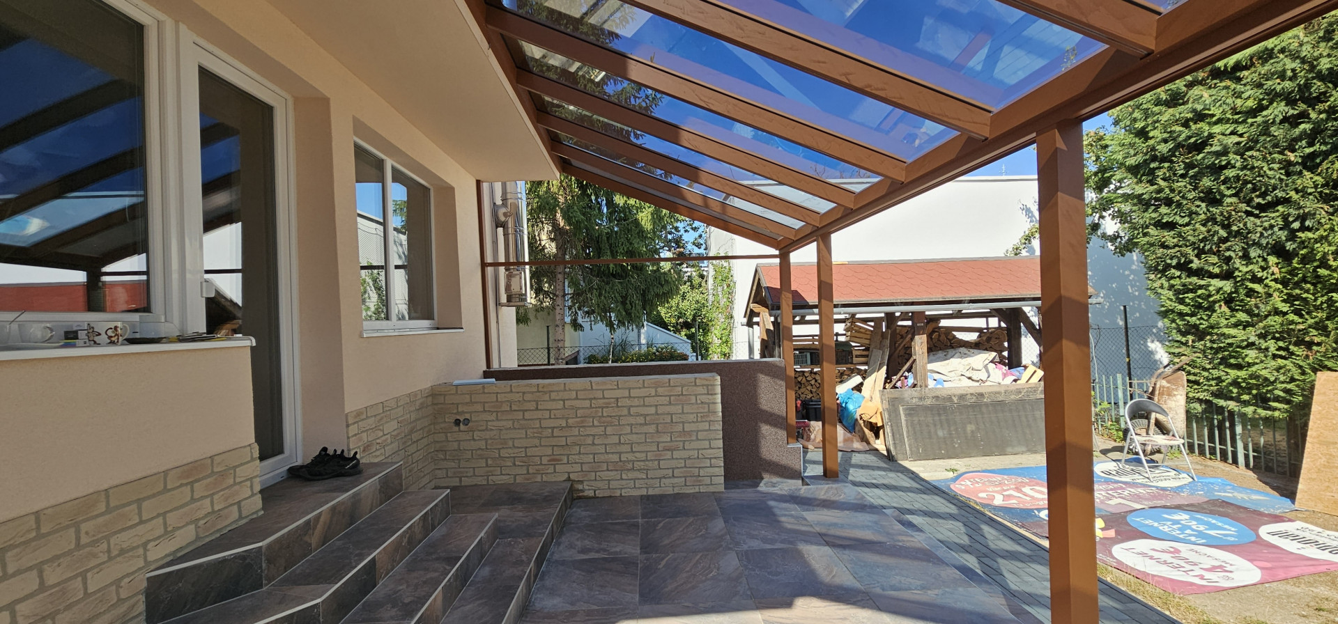Aluminiowa pergola o wykończeniu imitującym drewno, ze szklanym dachem, zamontowana na tarasie domu jednorodzinnego.