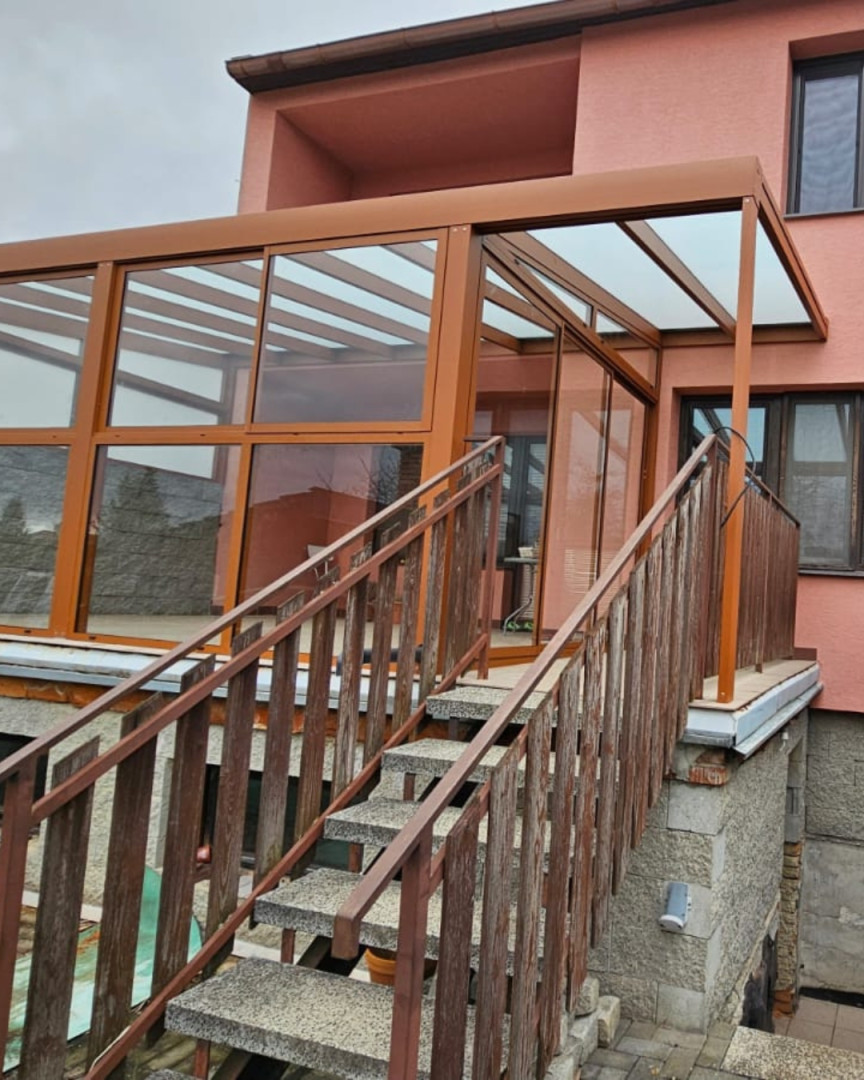 Szklana pergola z aluminium w kolorze brązowym, zamontowana na tarasie domu z klatką schodową