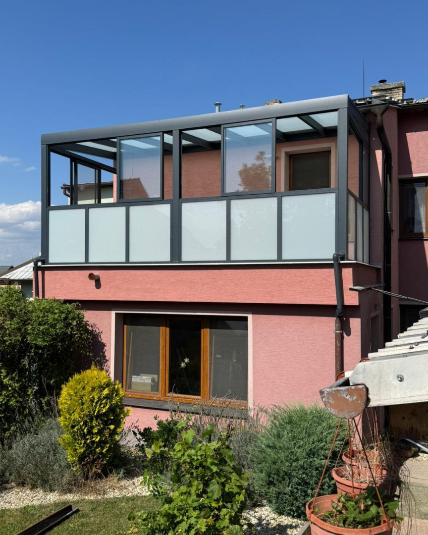 Nowoczesna przeszklona pergola z ciemnoszarą aluminiową ramą i mlecznym szkłem na balkonie domu jednorodzinnego.